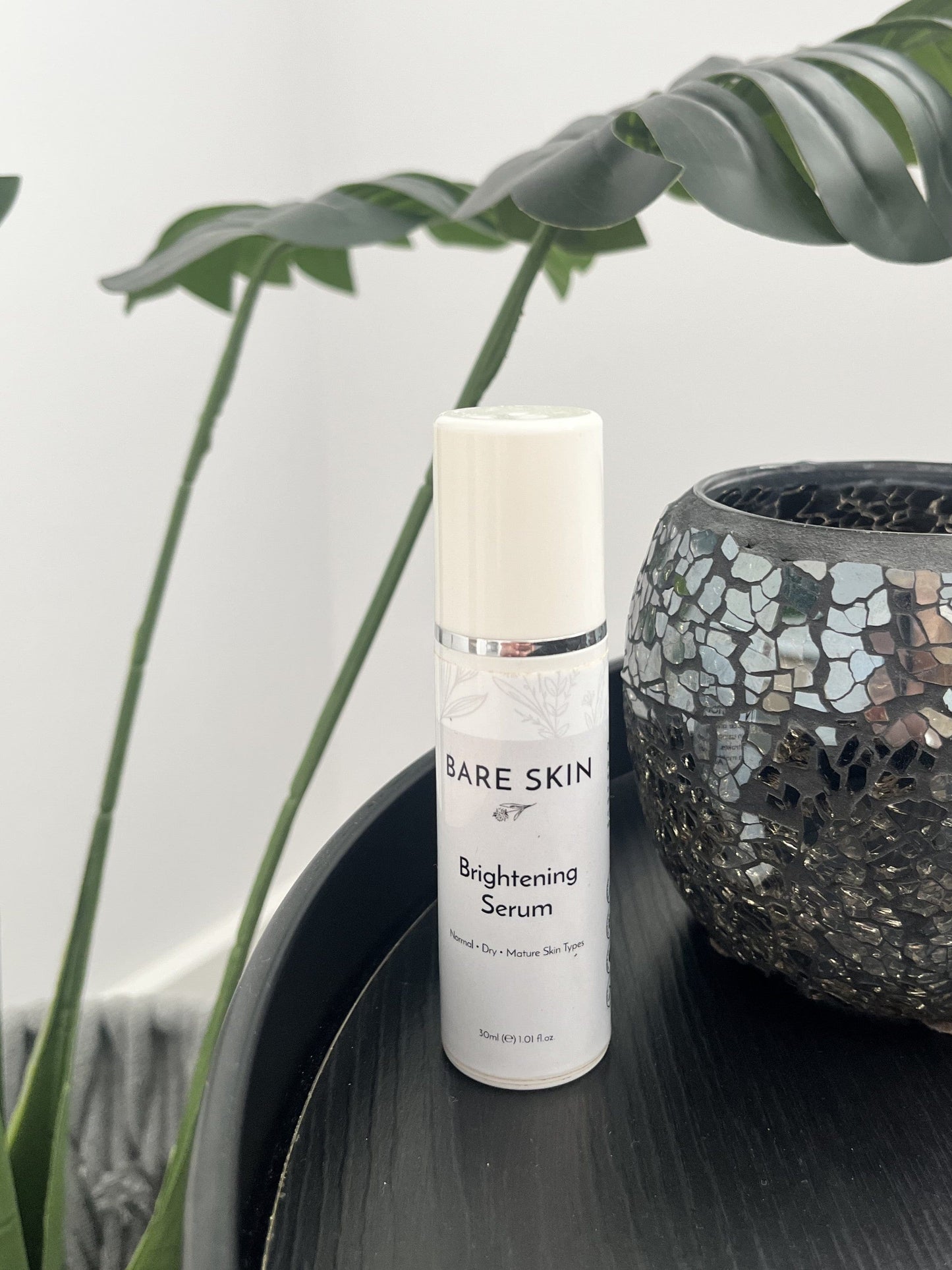 Bare Skin Brightening Serum bottle on a dark table with green plant in background, skincare for radiant even skin tone.