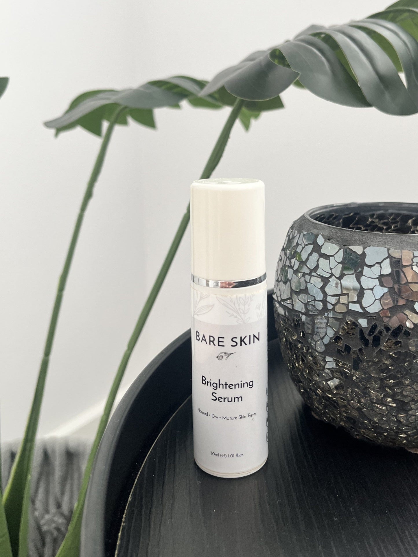 Bare Skin Brightening Serum bottle on a dark table with green plant in background, skincare for radiant even skin tone.