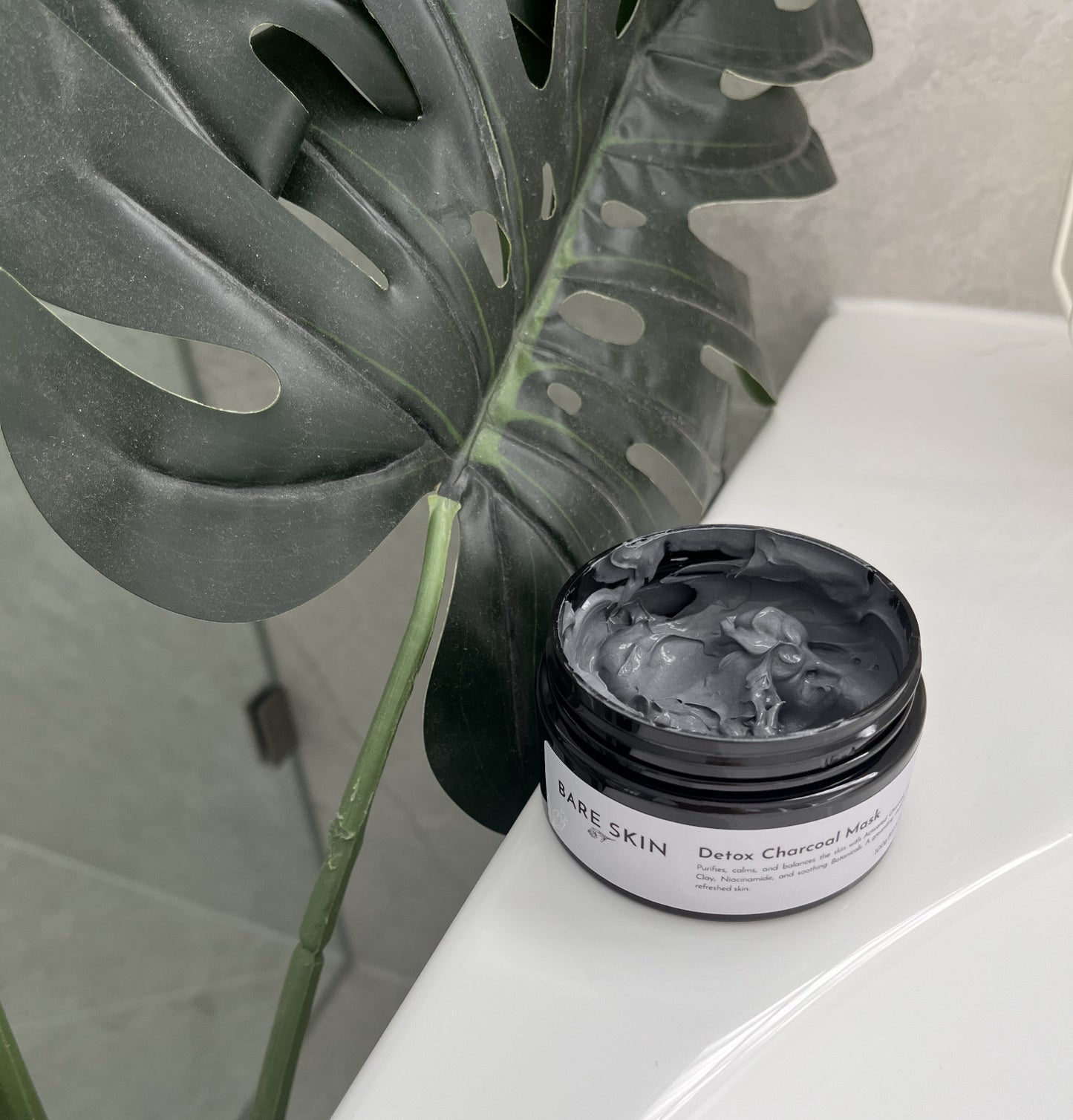 Jar of Bare Skin Detox Charcoal Mask on a bathroom counter with a plant in the background
