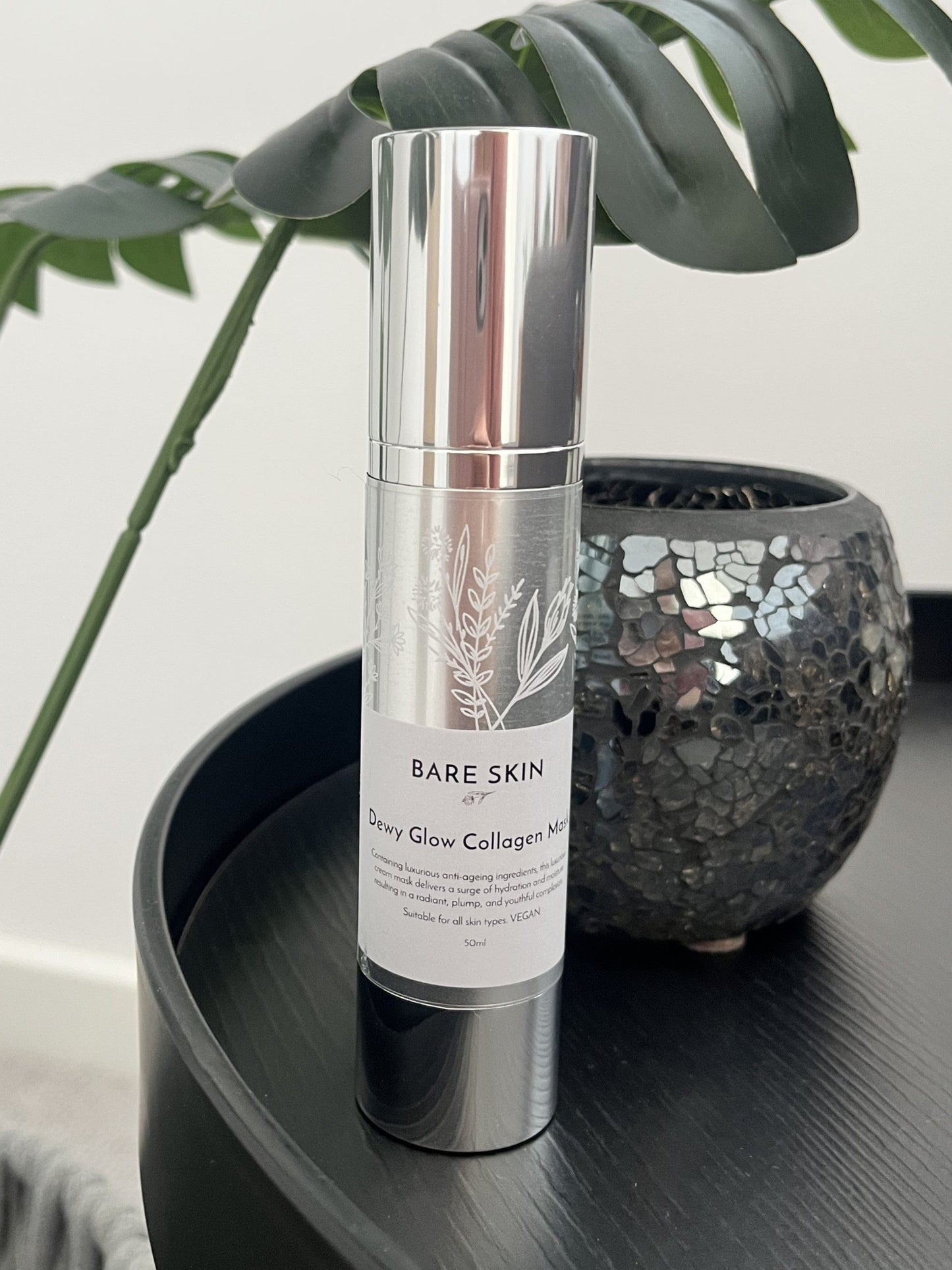 Bare Skin Dewy Collagen Mask bottle on dark round table with plant and mosaic vase in background, hydrating and firming overnight cream mask.