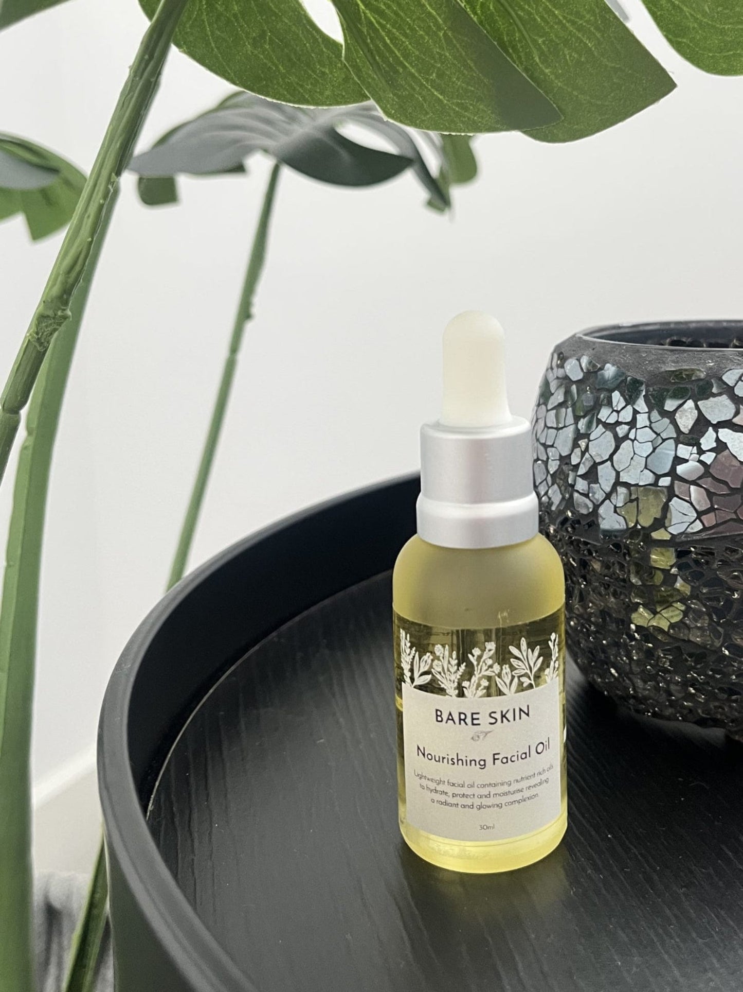 Bare Skin Nourishing Facial Oil bottle on a dark surface with green leaves in the background