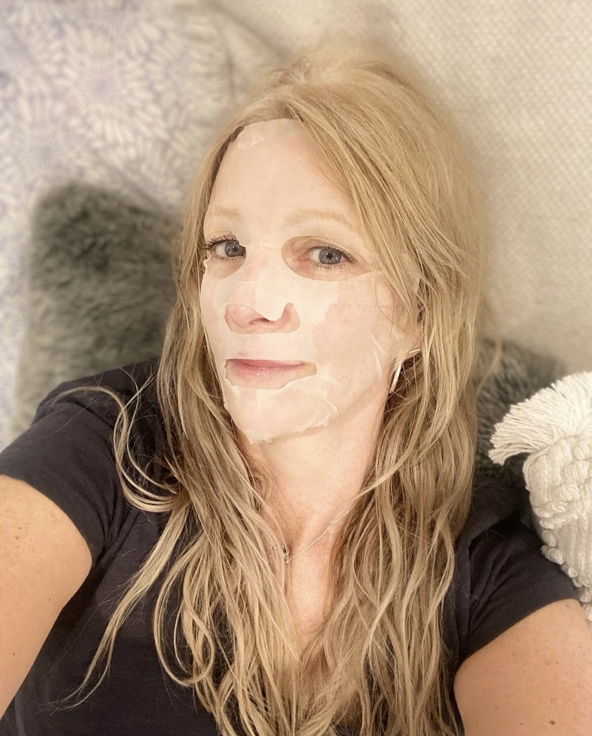 Woman laying on grey and white textured surface wearing Bare Skin Age Defying Sheet Mask, hydrating firming anti-aging skincare.