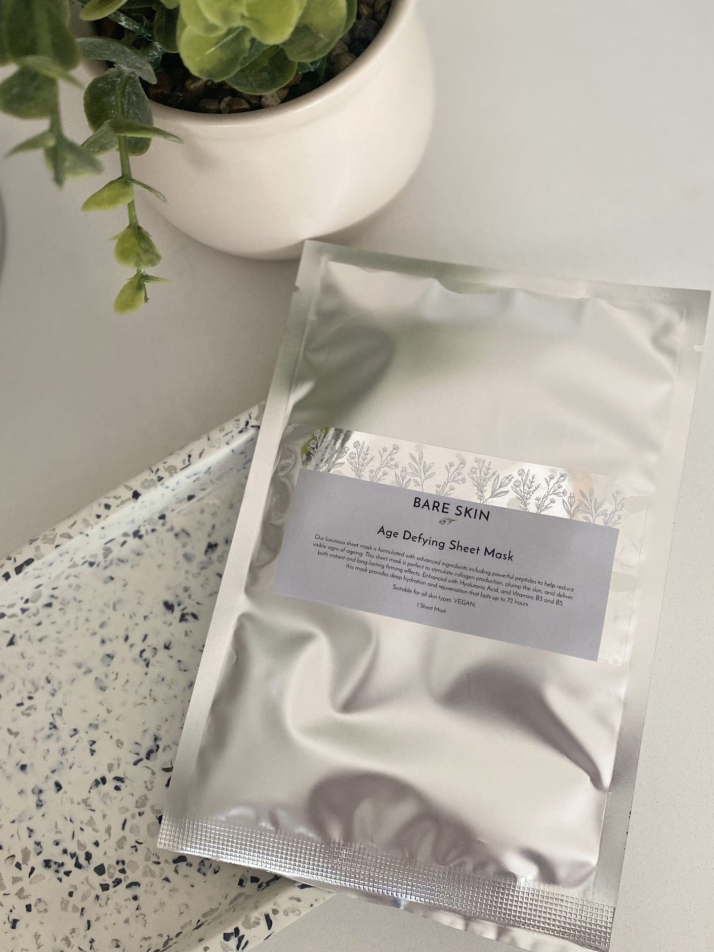 Bare Skin Age Defying Sheet Mask on speckled ceramic tray with cactus plant in background, hydrating firming anti-aging treatment.