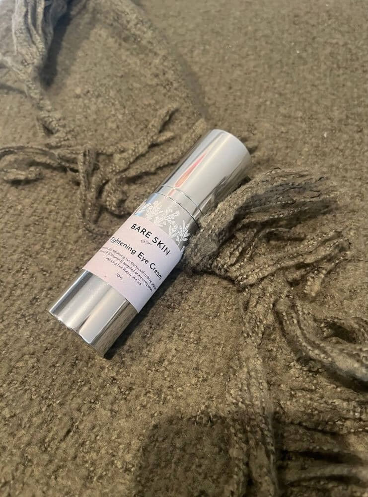 A silver tube of BARE SKIN Tightening Eye Cream resting on a textured, fringed brown knit blanket.