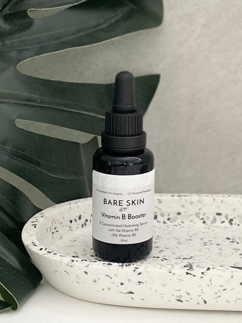Bare Skin Vitamin B Booster serum with Niacinamide and Hyaluronic Acid bottle on speckled tray with green leaves in background, hydration and barrier support.