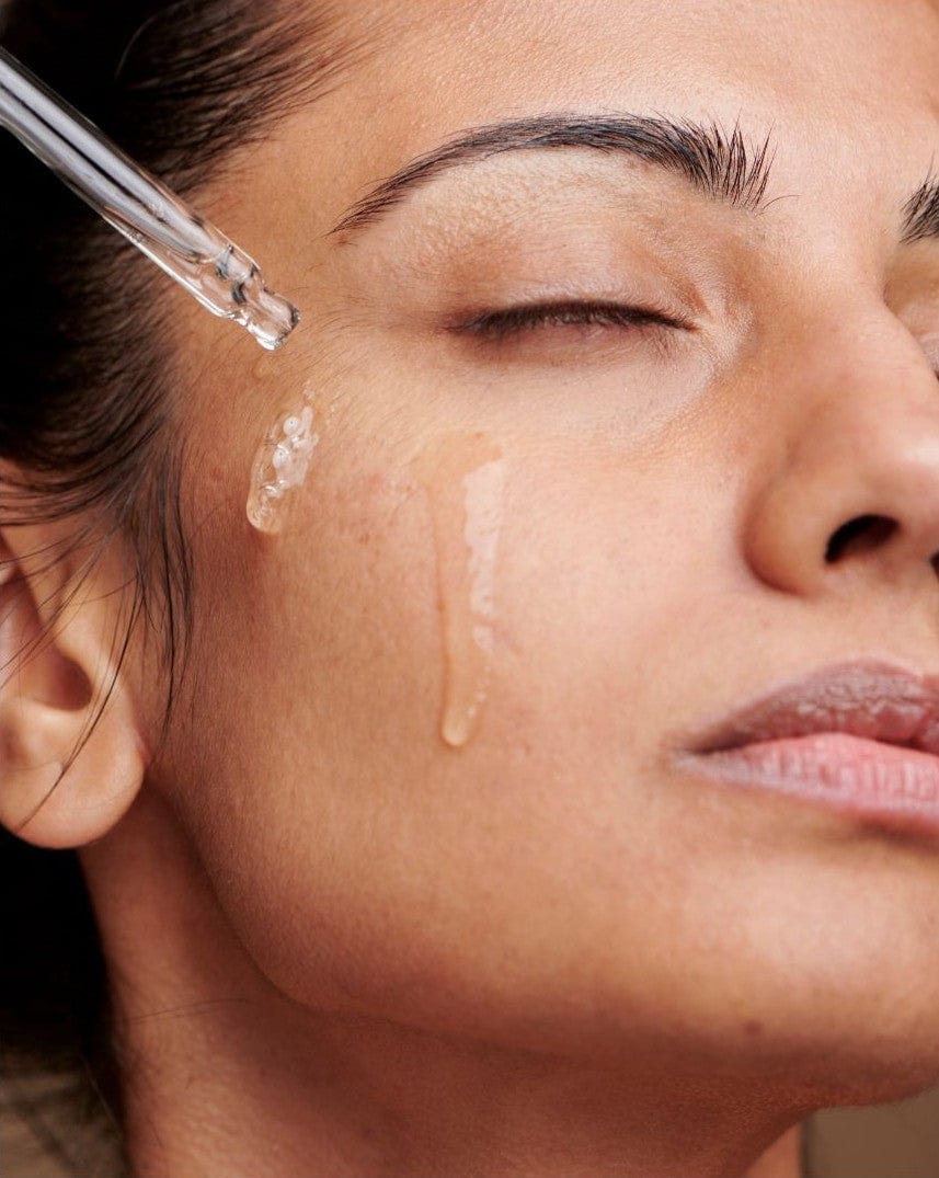 Close-up of woman applying Bare Skin Vitamin B Booster serum with Niacinamide and Hyaluronic Acid dropper to cheek, hydrating barrier-repair serum.