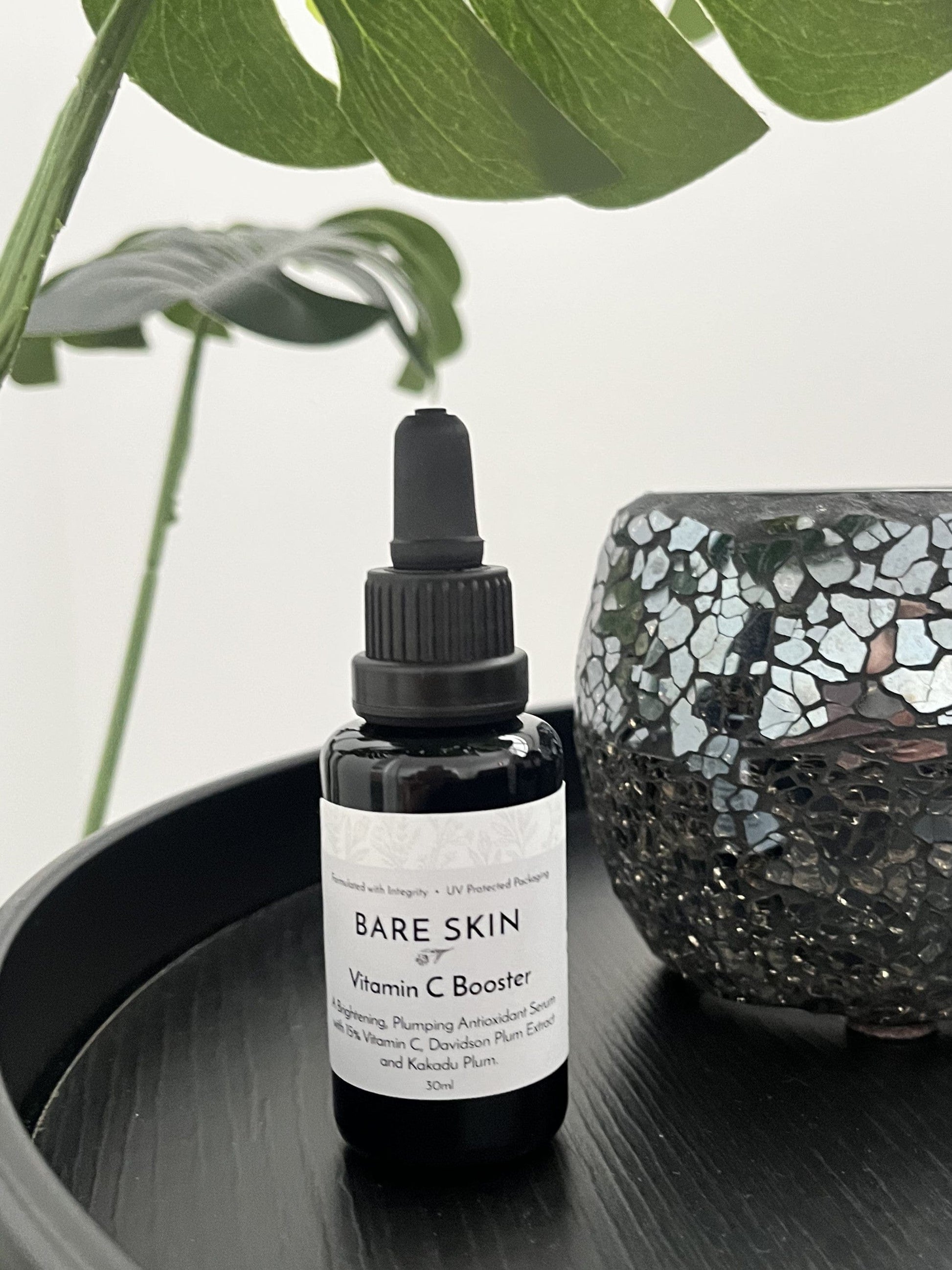 Bare Skin Vitamin C Booster brightening antioxidant serum with Kakadu Plum bottle on black round table with plant and mosaic vase in background.