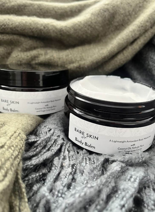Two jars of Bare Skin Body Balm arranged on a textured grey knit blanket with a green knit throw in the background. One jar is open, showing white balm inside, and the other is closed in the background.