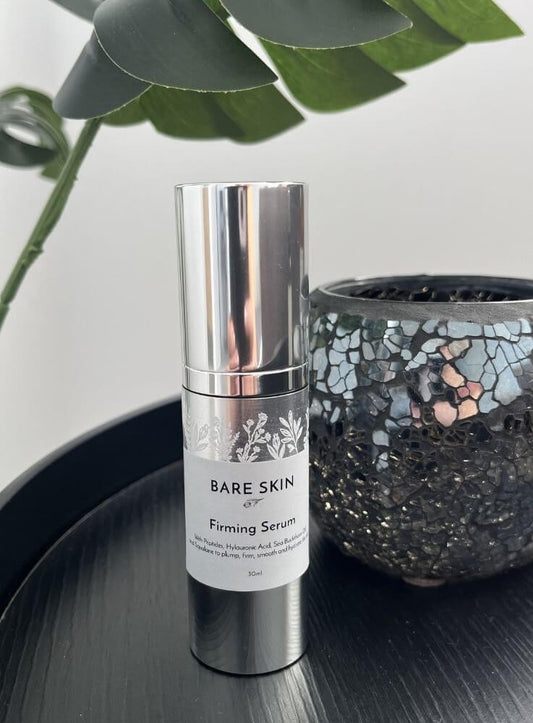 A silver cylindrical bottle of Bare Skin Firming Serum with a white label, standing on a black tray next to a mosaic glass container and green leaves in the background.