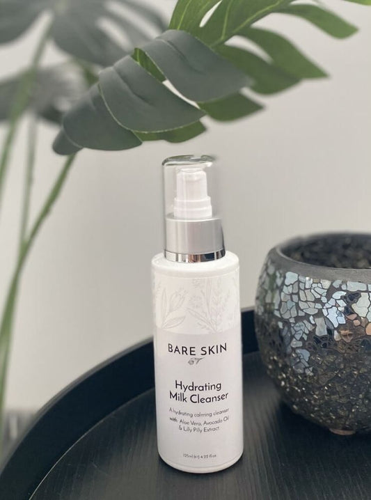 Bare Skin Hydrating Milk Cleanser bottle on a dark surface with green leaves in the background