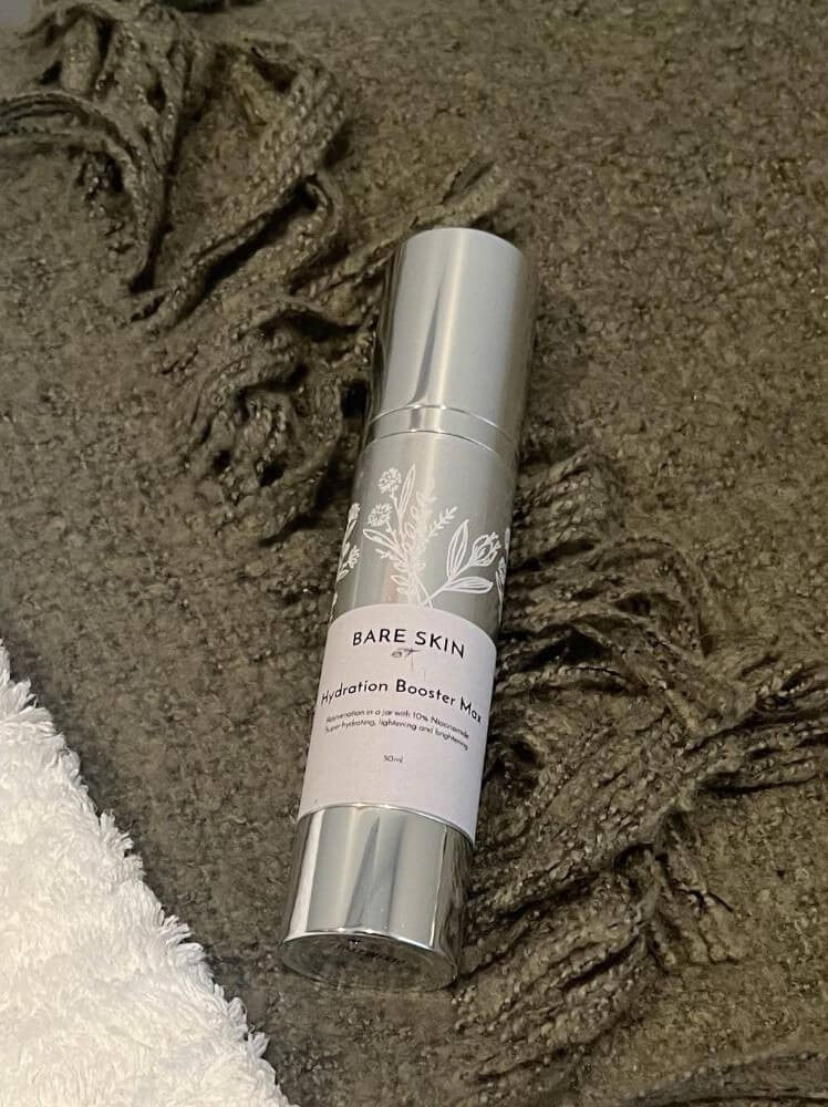 A silver cylindrical cosmetic bottle Bare Skin Hydration Booster Max resting on a textured green blanket with fringe, next to a white towel.