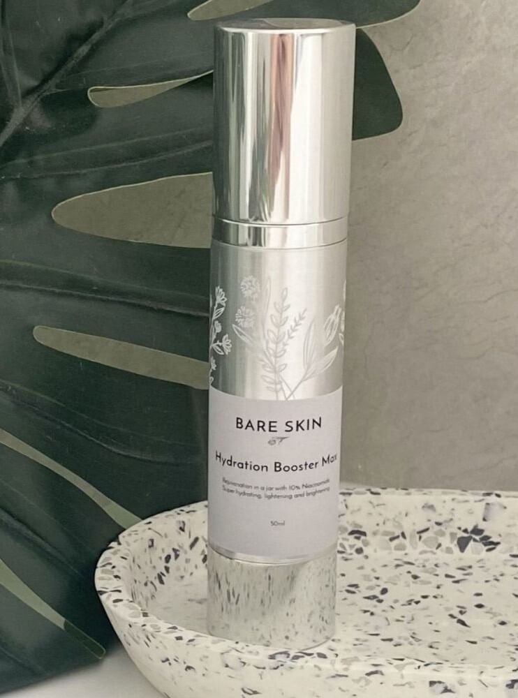 A silver cylindrical skincare Bare Skin Hydration Booster Max with a white label and botanical print, set on a speckled white tray with a green leafy background.