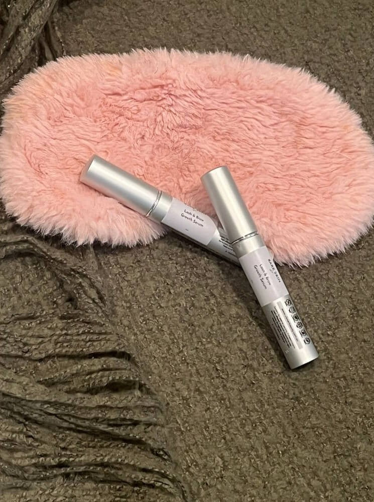 Two silver cosmetic tubes Bare Skin Lash & Brow Serum resting on a pink faux-fur oval cushion over a textured brown knit blanket.