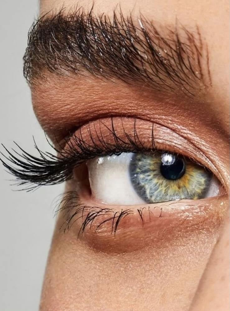 Extreme close-up of a human eye wearing a realistic blue-gray iris contact lens with a yellow-gold ring and long black eyelashes.