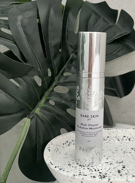 A silver cylindrical bottle of Bare Skin Multi-Vitamin Hydration Moisturiser standing on a white speckled tray, with a large dark green monstera leaf behind it.