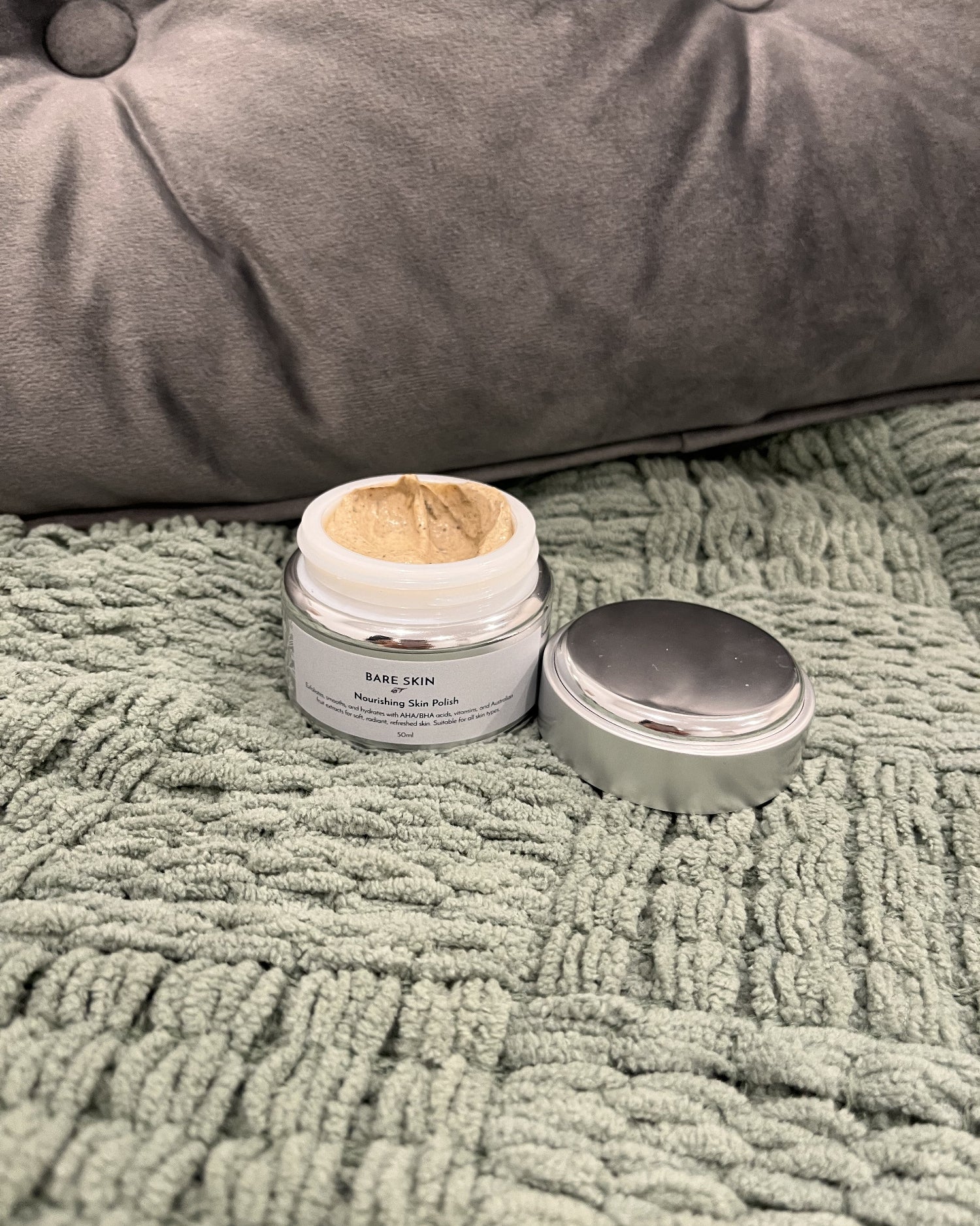An open jar of Bare Skin Nourishing Polish with its silver lid placed beside it on a textured sage green blanket, with a grey upholstered cushion in the background.