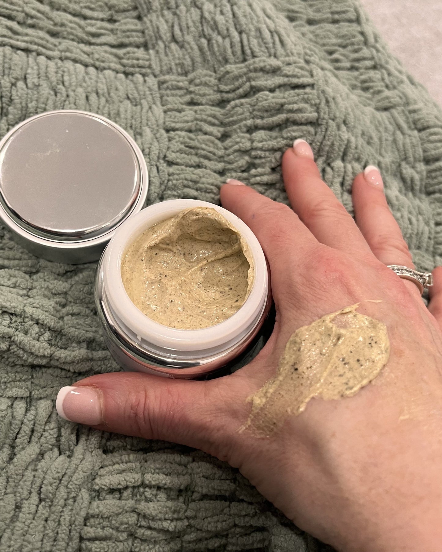 An open jar of Bare Skin Nourishing Skin Polish sitting on a ribbed sage green blanket, with a hand holding the jar and a smear of the product visible on the back of the hand.