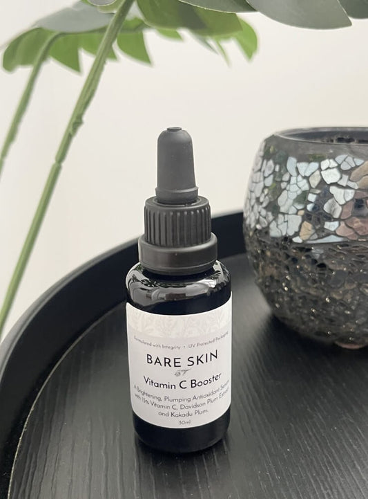 Bare Skin Vitamin C Booster brightening antioxidant serum with Kakadu Plum bottle on black round table with plant and mosaic vase in background.